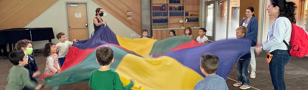 A group of children and teachers stand in a circle around a multicolored play parachute. They are laughing and waving the parachute up and down