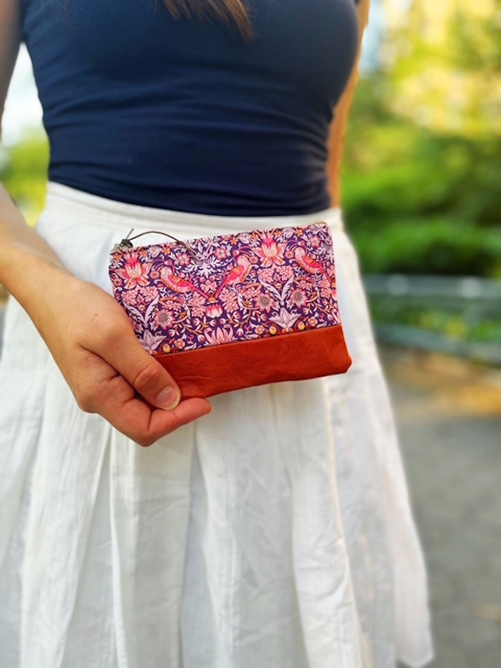 A woman holding a coin pouch with a strawberry thief design and leather accented outdoors in the park