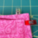 A quilt block with a pink binding partially folded to the back. The binding is folded around the corner, held in place with a yellow clip and a red clip.