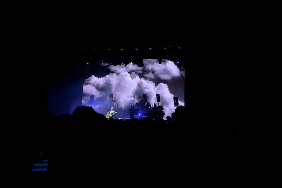 A woman on the left side of a stage holds a guitar and stands in front of a projection of a cloud.