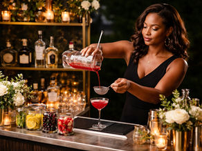 Bartender in a black dress pouring red cocktail through a strainer into a glass. Bar adorned with roses and candles, warm ambiance.