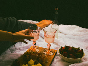 Two hands holding pink wine glasses on a bed with a charcuterie board and strawberries. Soft lighting, relaxed, cozy atmosphere.