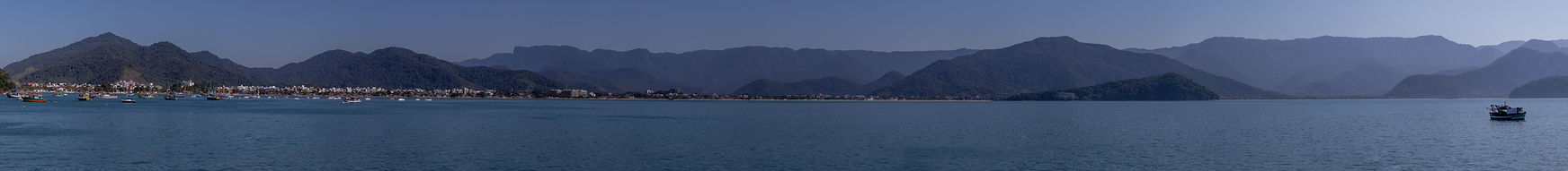 Vista panorâmica da baía de Ubatuba com águas calmas e azuladas, cercada pela Serra do Mar ao fundo. Na orla, vê-se a cidade litorânea de Ubatuba com diversas embarcações pequenas espalhadas pelo mar, incluindo um barco em destaque à direita da imagem. O céu está limpo, com tons suaves de azul.