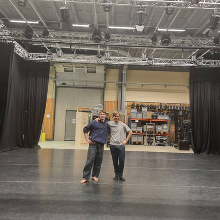 Två personer i träningskläder står på en scen med svart dansgolv och väggar och teknisk utrystning bakom.