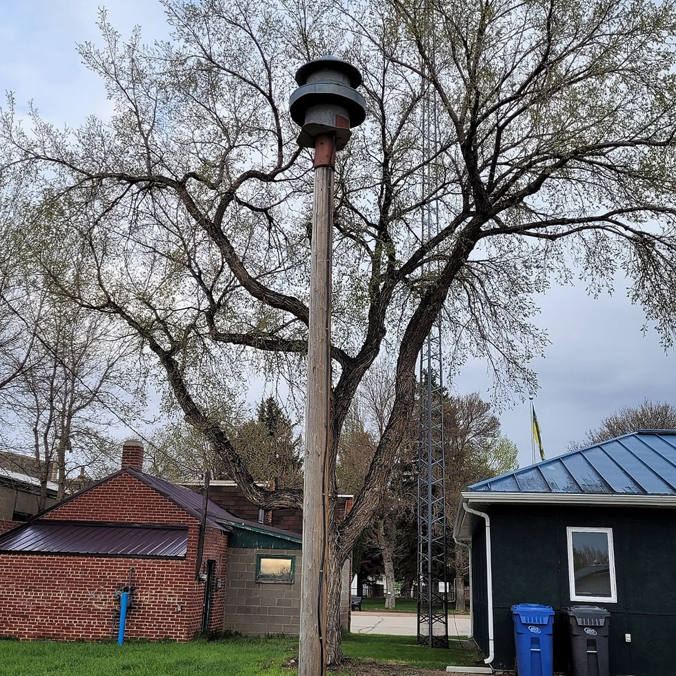 Gull Lake siren mounted on a wooden pole amid trees beside a brick building and blue-roofed structure, with blue bins and a cloudy sky overhead.