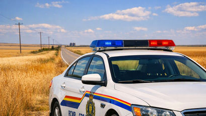 White RCMP police car with flashing red and blue lights on a rural road through golden fields under a blue sky.