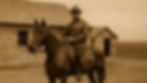 Man in uniform on a horse in front of wooden buildings in sepia tone. The setting is open landscape, evoking a historical mood.