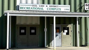 Green building with double doors, sign reads "Gull Lake & District Recreational Complex (1984)." Sunlit, simple, and functional entrance.