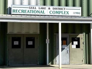 Green building with double doors, sign reads "Gull Lake & District Recreational Complex (1984)." Sunlit, simple, and functional entrance.