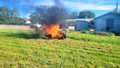 Lawn tractor engulfed in flames and black smoke in a grassy yard near houses on a sunny day.