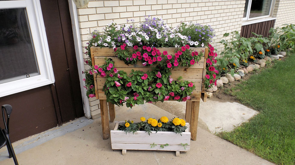 Wooden flower planters overflowing with pink, white, purple, and yellow blooms beside a brick house and patio garden