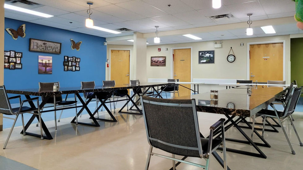 Dining room with black tables, gray chairs, blue wall with photos, and a "Believe" sign. Clock on a beige wall, doors closed.