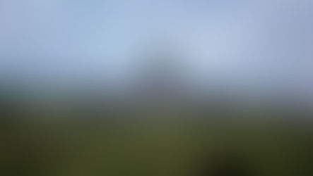 Grain silos and storage tanks on a farm under a cloudy sky, surrounded by green fields with wind turbines in the background.