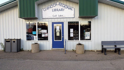 Facade of Chinook Regional Library, Gull Lake. Blue door, white walls, green awning. Benches, trash bins, "OPEN" sign on window.