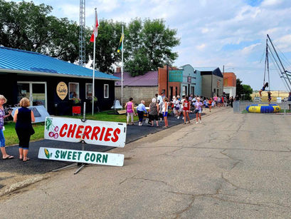 Crowd gathering near a building with a blue roof, signs for "Cherries" and "Sweet Corn" visible. Carnival ride to the right, cloudy sky above.