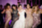 BusyBrides Wedding with ladies in elegant dresses dance joyfully in a festive setting with purple lighting. The mood is lively and celebratory.