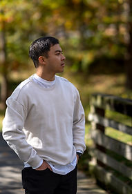Portrait of Matthew wearing a light sweatshirt, standing near a wooden fence under fall-colored trees.