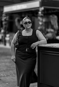Black and white portrait of Natalie smiling while leaning on a ledge in a casual city setting.