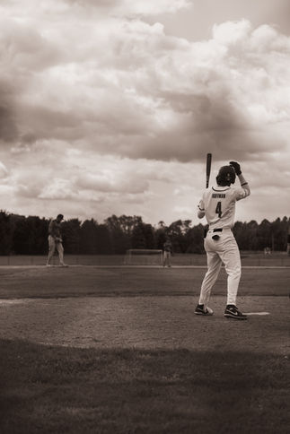 A player named Hofman prepares to bat under dramatic clouds, standing near home plate as the pitcher readies on the mound in the distance.