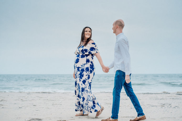expecting woman smiles back at husband as they walk along the beach shore