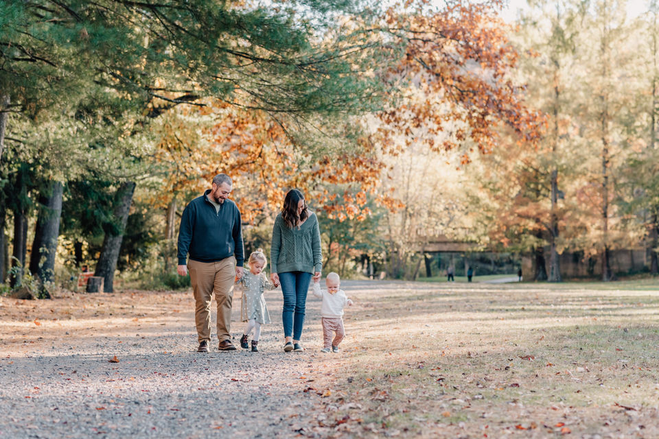 young family holds hands and walks down trail surrounded by leaves