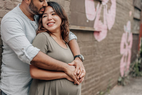 man hugs smiling pregnant wife from behind in front of a floral brick wall