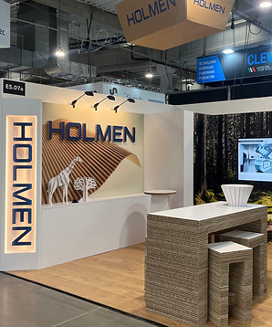 Oversigtsbillede af Holmen-standen med belyst vertikalt skilt, receptionsdisk og møbler i re-board