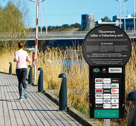 TiksPac miljöstation i Falkenberg med gratis skräppåsar och lokala företagslogotyper. Stationen är placerad vid promenadstråk och används för att hålla kommunen ren, här passerar en kvinna som joggar.