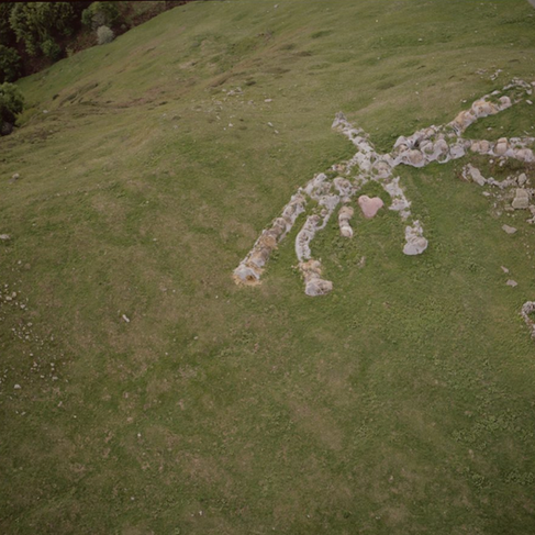 Veduta zenitale dei resti dell'installazione "Hase" anni dopo la sua creazione. Il corpo del coniglio è quasi completamente decomposto e sbiadito in un grigio terroso, mimetizzandosi con il terreno e le rocce circostanti. Al centro della sagoma ormai consunta è ancora visibile un unico elemento residuo di colore rosa acceso a forma di cuore, che rappresenta le interiora stilizzate del pupazzo.