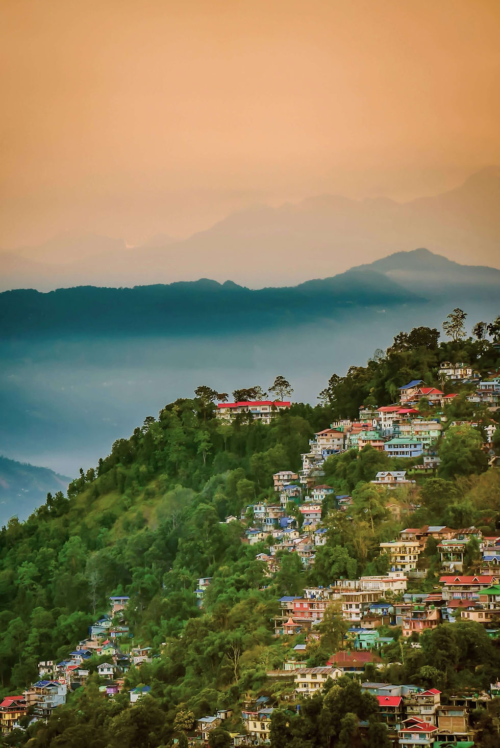 A breathtaking view of Kalimpong, a charming hill town set against the backdrop of majestic mountains. Nestled in the lush green landscapes of West Bengal, India, Kalimpong’s colorful houses dot the slopes, creating a striking contrast against the dense foliage. Layers of misty blue hills fade into the horizon beneath a warm, golden sky, adding to the town’s ethereal beauty. Captured at sunset, this serene scene perfectly encapsulates the tranquility and allure of this Himalayan gem.