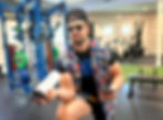 Man in sunglasses and floral vest crouches in a gym, holding a shaker bottle and phone beside blue weight racks.