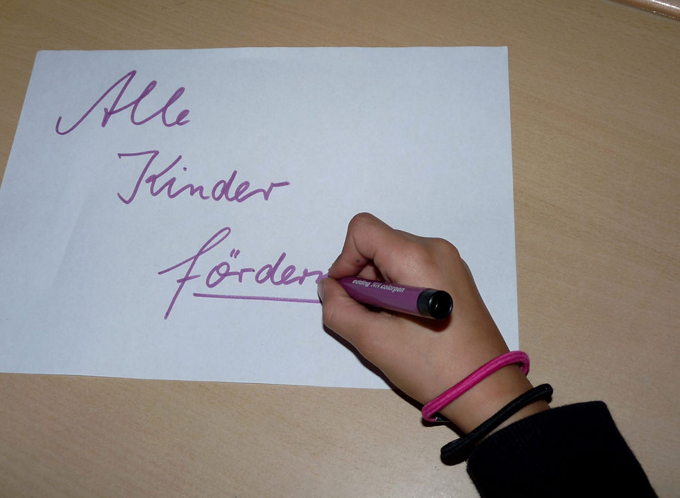 Kinderhand schreibt den Schriftzug Alle Kinder foerdern mit violettem Stift auf Papier