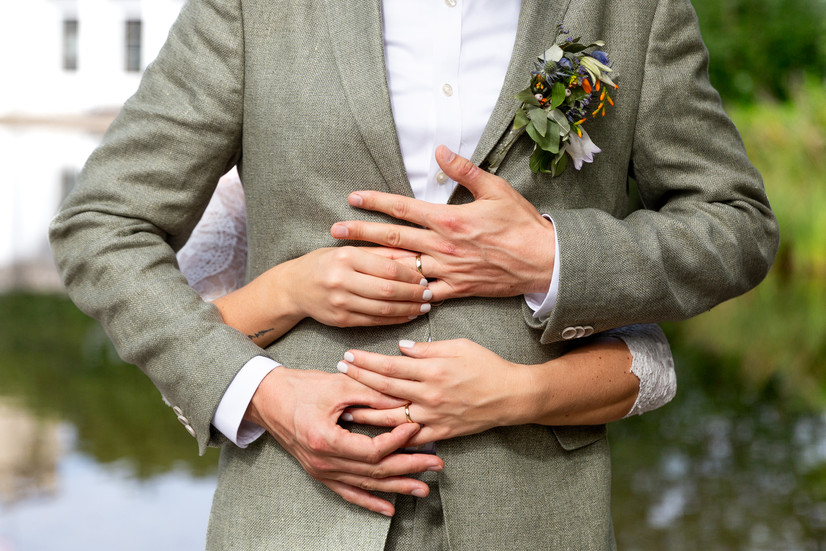 242 Das Brautpaar befindet sich im Park vor dem Landratsamt Schöneberg in Berlin. Vom wunderschön verschwommenen Hintergrund ist nur wenig zu erkennen; lediglich ein kleiner, dunstiger Teich. Die beiden halten die Eheringe des jeweils anderen mit verschränkten Händen. Nur ihre Oberkörper sind sichtbar.