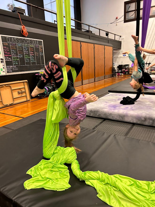 Two kids practicing aerial silk acrobatics indoors. One hangs from bright green fabric, another in teal. Chalkboard with "Class Schedule" visible.
