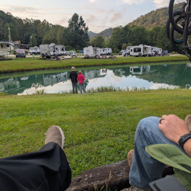 Two people stand by a pond reflecting green RVs and trees in a park. Camping chairs and relaxed feet are in the foreground.