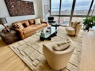New Jersey. Living room with a leather sofa, sculptural lounge chairs, a glass coffee table, layered neutral textures, and city views through large windows.