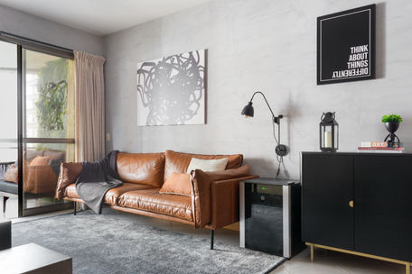 Residential living room in Rio de Janeiro with leather sofa, abstract wall art, black storage cabinet, wall-mounted reading lamp, and neutral layered textiles