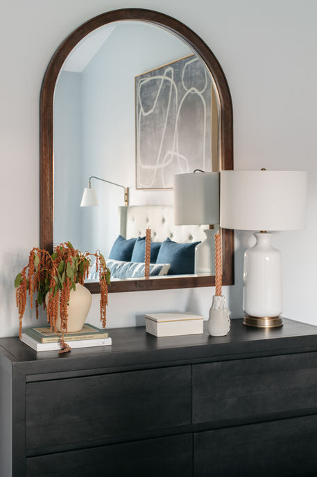 Primary bedroom dresser styling with dark wood dresser, arched wood-framed mirror, white table lamps, ceramic vases, and soft blue accents reflected in the mirror