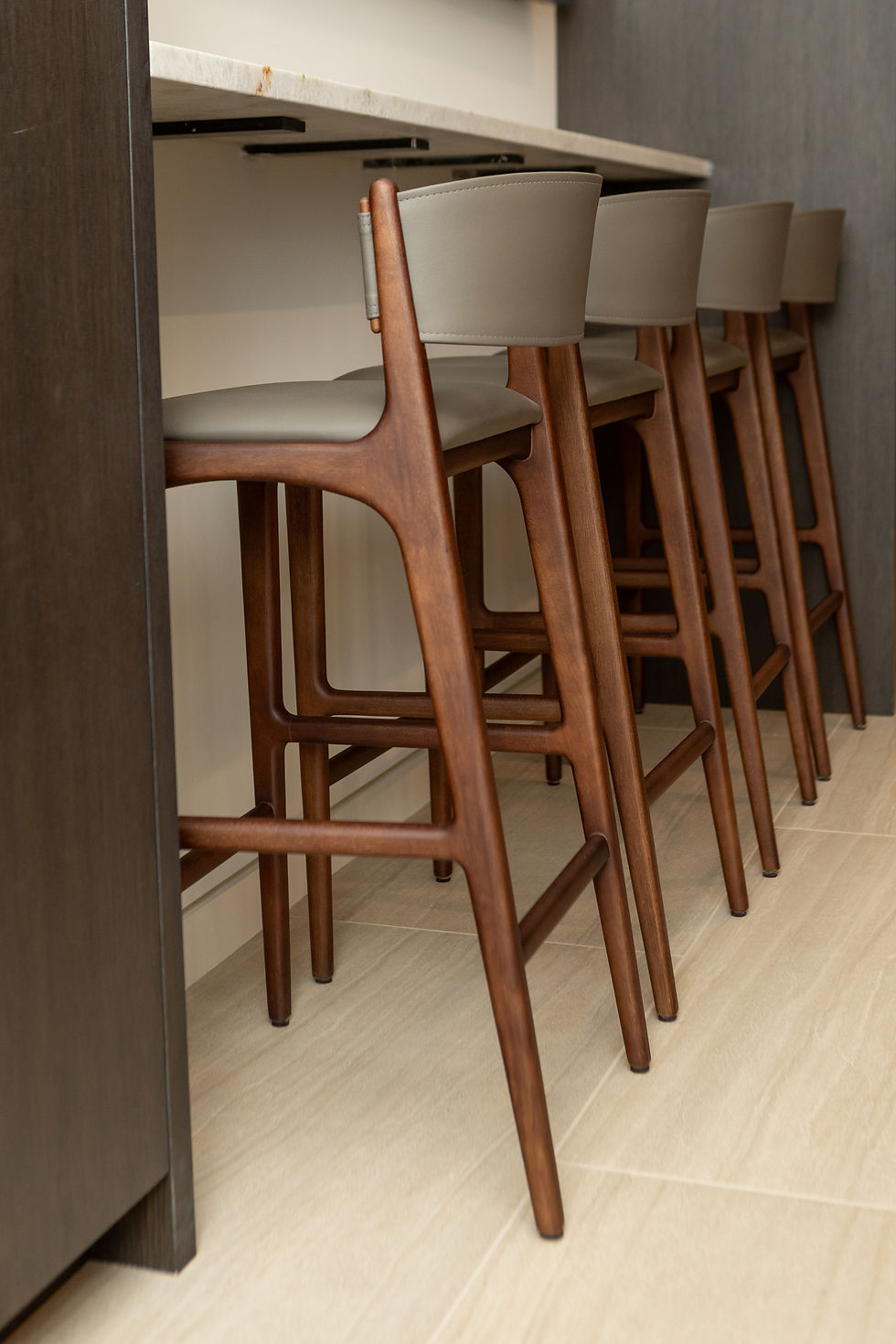 A close-up side view of three Estúdio Bola Helga bar stools tucked under a light stone countertop. The stools feature a curved solid wood frame with a warm walnut finish and taupe leather upholstered seats and backrests, showcasing the seamless joinery and elegant profile.
