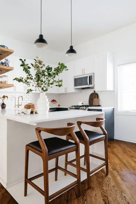 Bright kitchen in an Evanston, Illinois home featuring a white waterfall island with bar seating, warm wood stools, matte black pendant lights, white cabinetry, open wood shelving, and layered natural textures for a clean yet inviting feel.