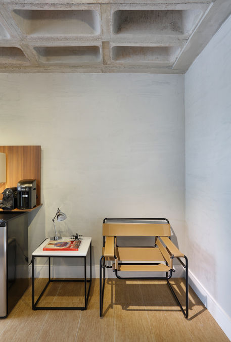 Commercial office design in Rio de Janeiro featuring a minimalist lounge area with leather accent chair, side table, task lamp, and exposed concrete ceiling