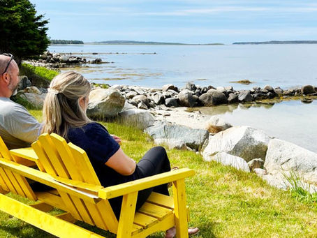 Two people sit on yellow chairs, overlooking a rocky beach with calm water. The mood is peaceful, with greenery and a clear sky.