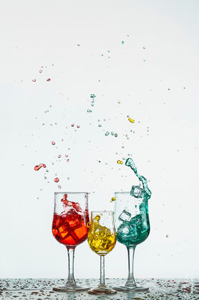 Drei Glaeser mit farbigen Fluessigkeiten im Splash-Moment vor hellem Hintergrund