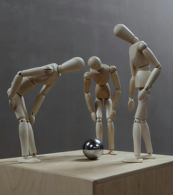 Three wooden figures leaning toward a small metallic ball on a platform