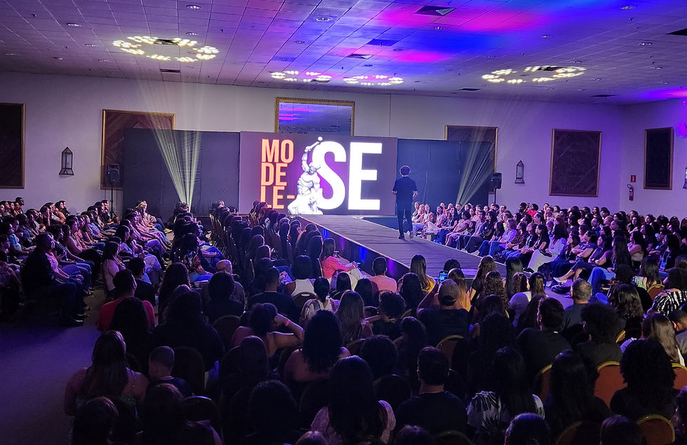 Pessoas assistem desfile de moda em salão com passarela iluminada. Tela ao fundo com "MODELO SE". Luzes roxas e azuis criam clima vibrante.
