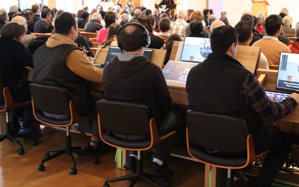 People in a crowded room focus on screens with headphones on. Wooden desks and various monitors are visible, creating a tech-oriented atmosphere.