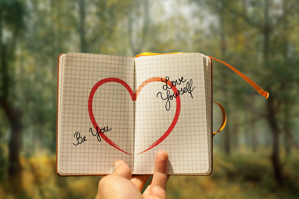Open notebook with a red heart and the words Be You, Love Yourself, held up against a blurred forest background.