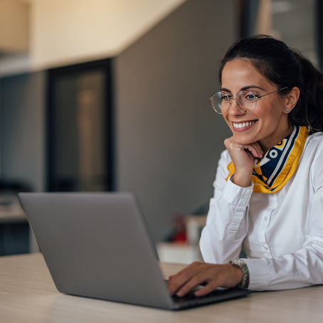 Mulher sorrindo enquanto trabalha em um laptop, sentada à mesa em um ambiente de escritório moderno e iluminado. Ela usa camisa branca, lenço amarelo e azul e óculos de grau.