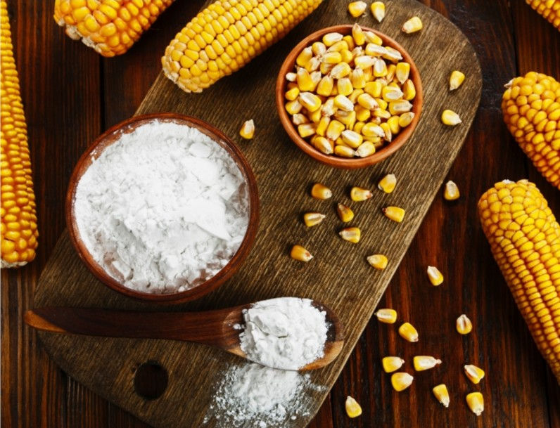 Corn cobs and kernels on a wooden board with bowls of cornstarch. A wooden spoon holds more starch, set on a rustic wood table.