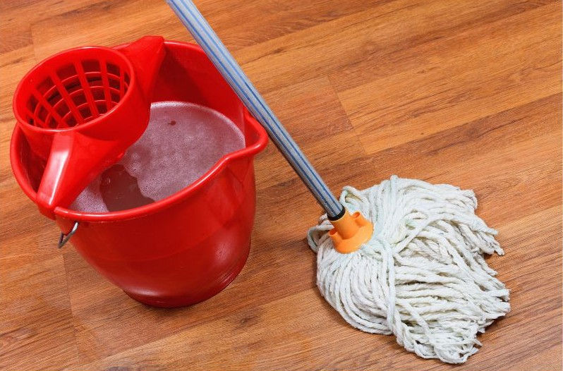 Red mop bucket with wringer and soapy water beside a white string mop on a wooden floor.